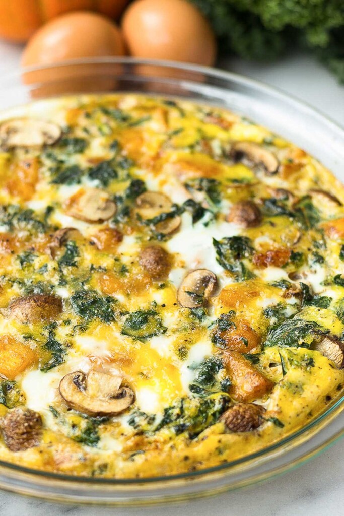 Crustless pumpkin quiche with veggies in a pie pan with eggs, pumpkin, and kale behind it.