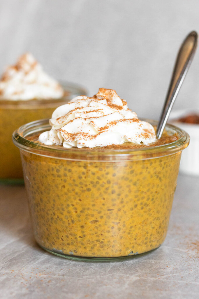 Healthy pumpkin chia pudding recipe in a jar topped with whipped cream and cinnamon. A spoon is coming out of the jar and another jar is behind it.