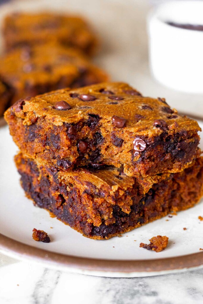 A small plate of two pumpkin bars stacked on top of each other with a bite taken out of the top one. Behind the plate is more jars and a small white dish.