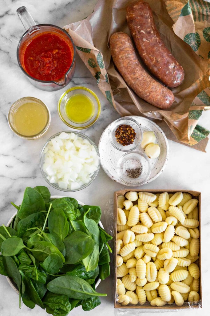 White marble counter with two raw links of Italian sausage, a small plate with two cloves of garlic, a bowl of red pepper flakes, and a bowl of salt and pepper, a container of gnocchi, a bowl of spinach, a bowl of diced onion, a bowl of olive oil, a jar of chicken broth, and a measuring cup of pasta sauce.