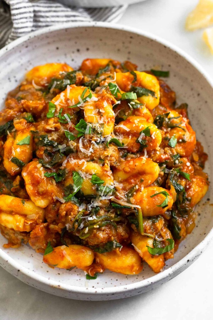 White plate with gnocchi & Italian sausage mixed with pasta sauce. It is garnished with basil and parmesan cheese.