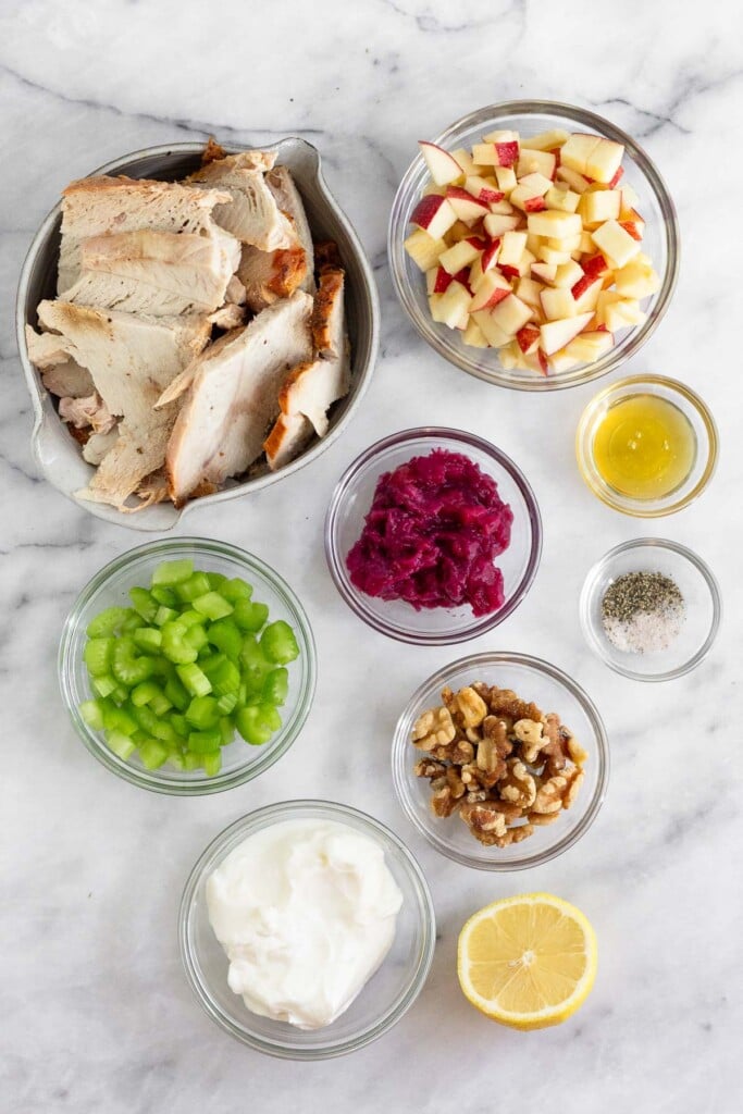White marble counter with a bowl of diced apples, a small bowl of honey, a bowl of salt and pepper, a bowl of cranberry sauce, a bowl of walnuts, half a lemon, a bowl of yogurt, a bowl of diced celery, and a bowl of slices of turkey.