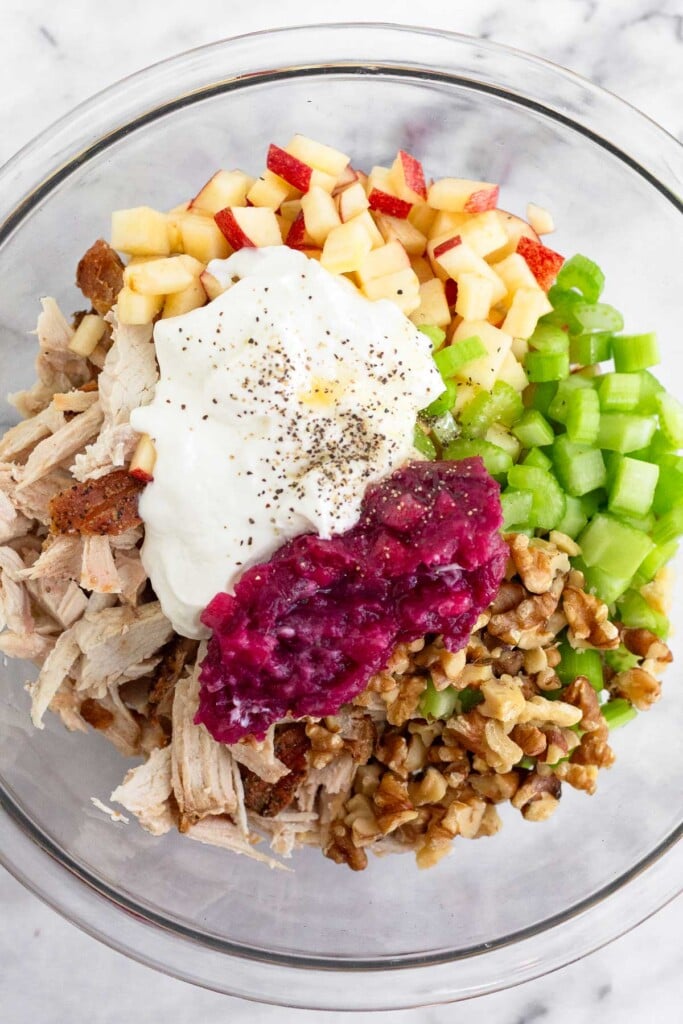 A large glass bowl with shredded turkey, diced apples, diced celery, chopped walnuts, yogurt, cranberry sauce, salt, and pepper.