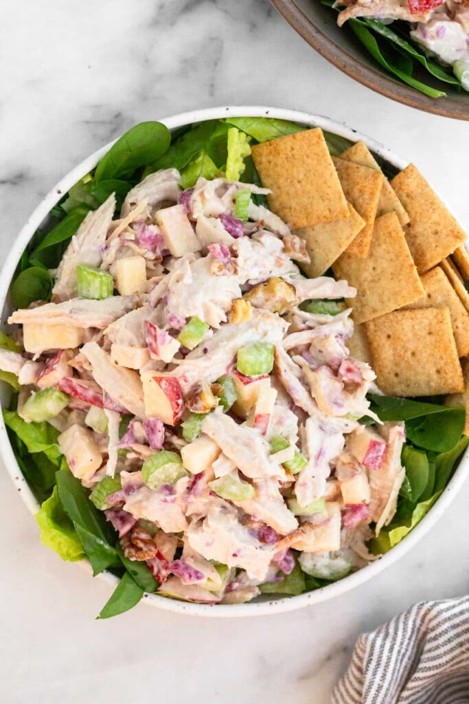 Turkey cranberry salad with apples, celery, and nuts overtop a bed of greens in a bowl. There are also some crackers in the bowl. Another bowl with more turkey salad is off to the side.