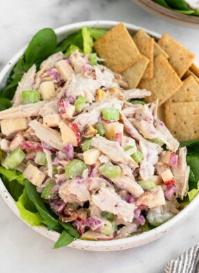 Leftover turkey salad with apples and celery in a bowl with some crackers overtop some greens. Behind the bowl is another bowl with similar contents.