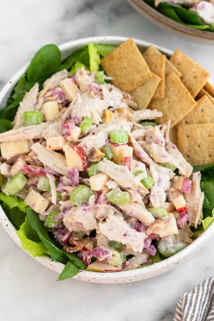 Leftover turkey salad with apples and celery in a bowl with some crackers overtop some greens. Behind the bowl is another bowl with similar contents.