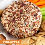 Pineapple cheese ball coated in chopped nuts on a dish surrounded by crackers, celery sticks, and carrots sticks.