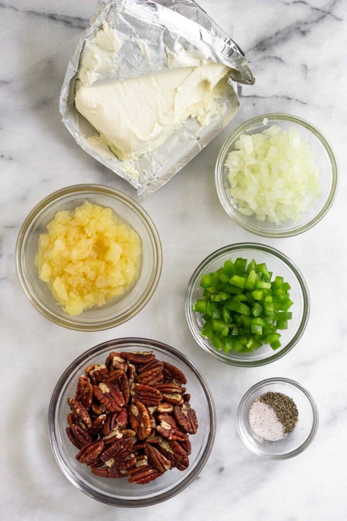 White marble counter with a package of cream cheese, a bowl of diced onions, a bowl of diced green peppers, a bowl of salt and pepper, a bowl of pecans and a bow of crushed pineapple.