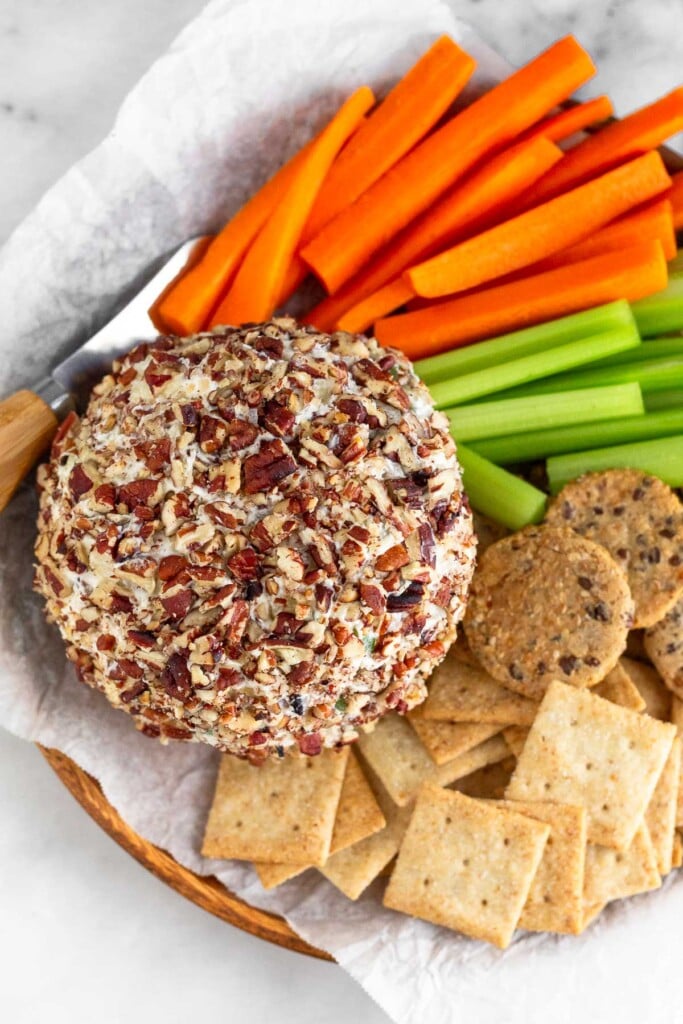 Overhead shot of a cheese ball coated in nuts with crackers, veggies, and a small cheese knife around it on a plate.