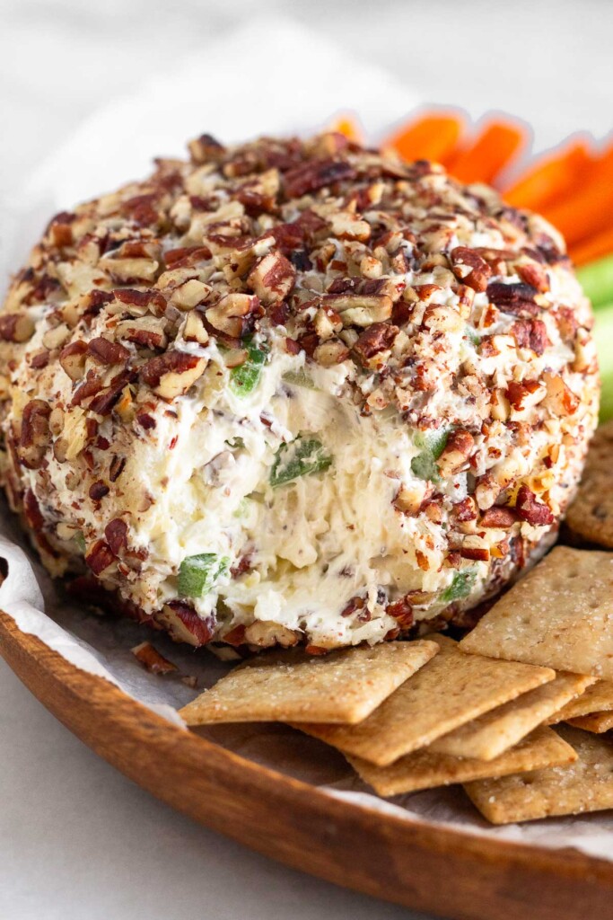 Cream cheese ball coated in chopped nuts on a plate surrounded by crackers and veggies. Some of the cheese ball has been eaten exposing the middle of it.