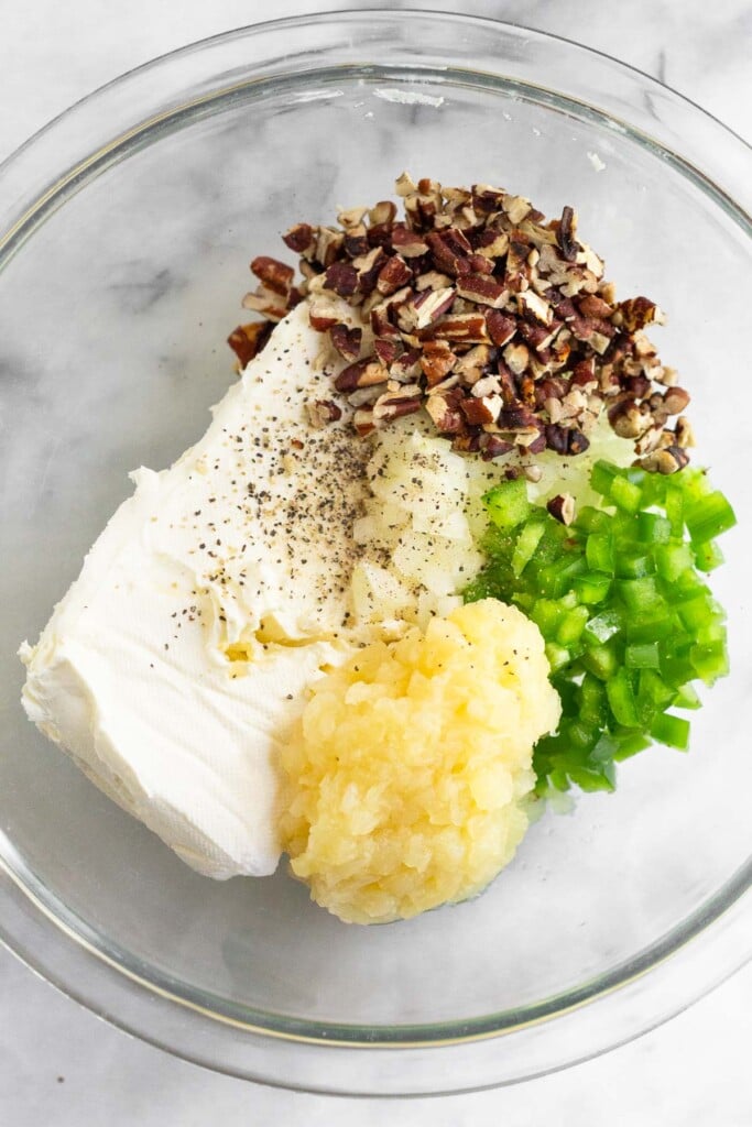 Large glass bowl filled with a brick of cream cheese, chopped nuts, diced onion, diced pepper, crushed pineapple, salt, and pepper.