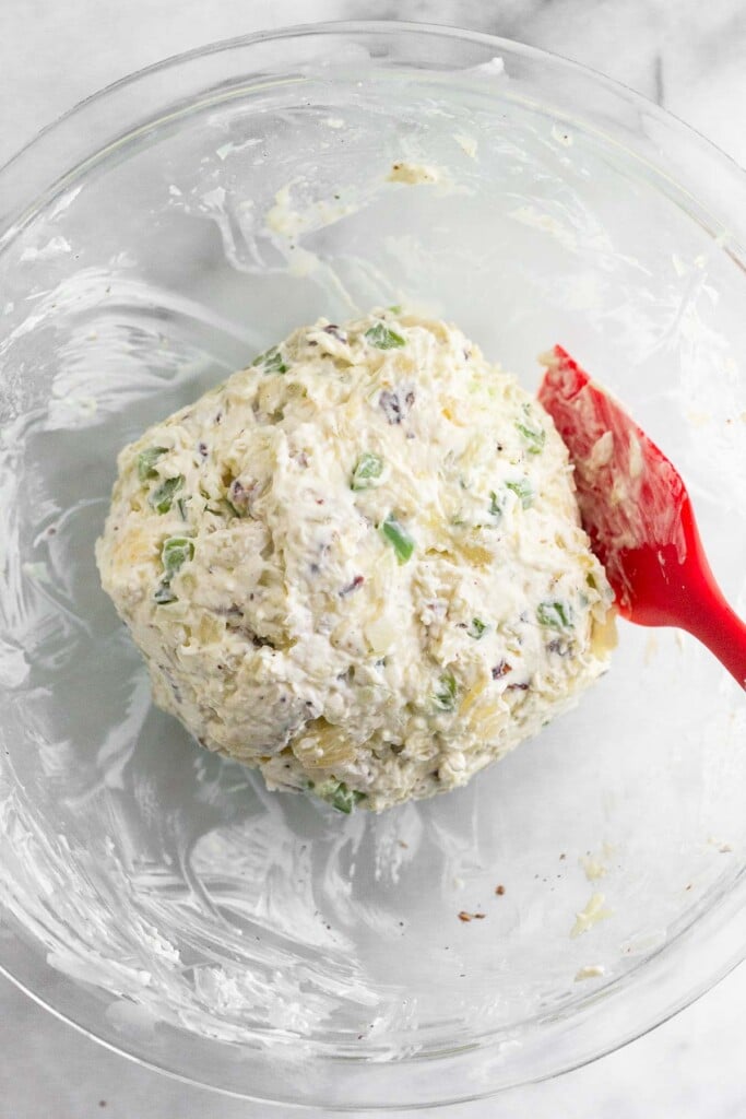 Pineapple cream cheese ball with peppers and nuts in a bowl after it has jut been formed in to a ball. A red spatula is also in the bowl.