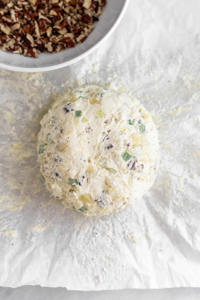 Cream cheese cheese ball on a piece of parchment paper. Next to it is a bowl of chopped nuts.