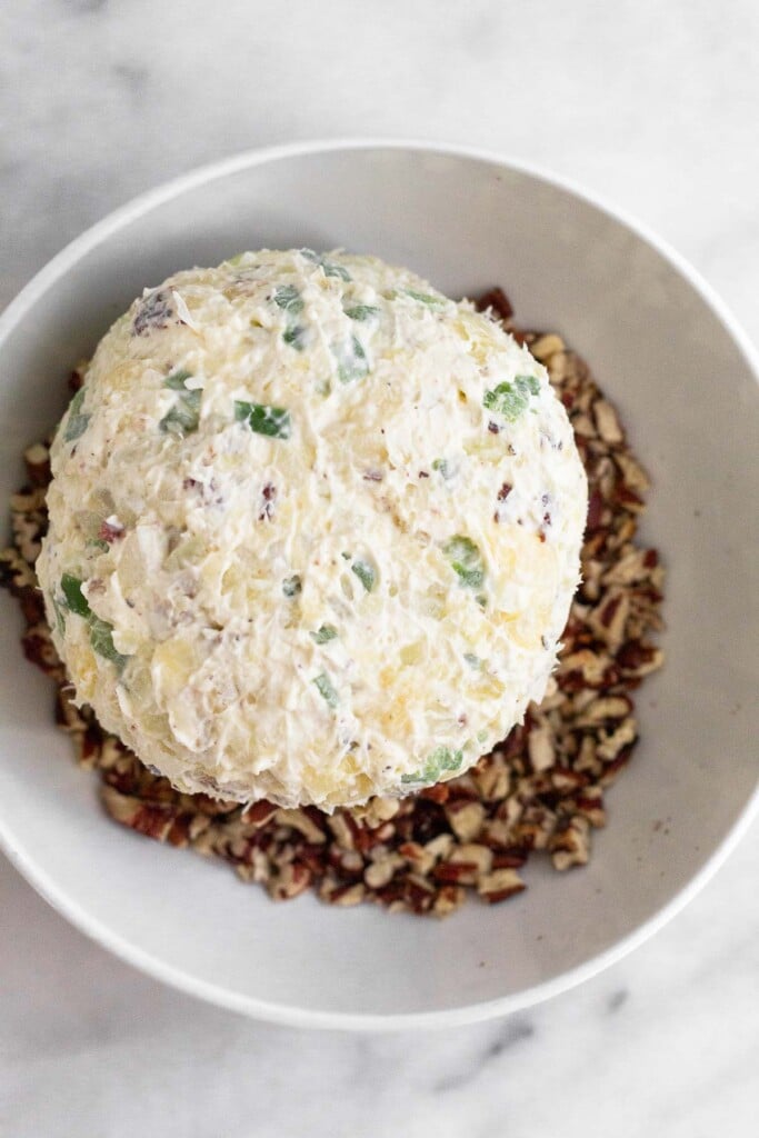 Cheese ball with pineapple, peppers, and nuts on top of a pile of chopped nuts in a bowl.