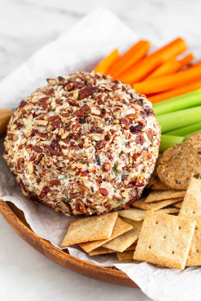 Pineapple cheese ball coated in chopped nuts on a dish surrounded by crackers, celery sticks, and carrots sticks.