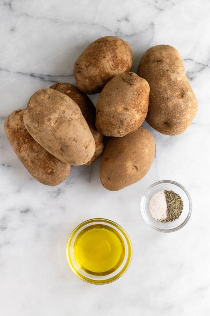 White marble counter with a pile of russet potatoes, a small bowl of salt and pepper, and a bowl of olive oil.