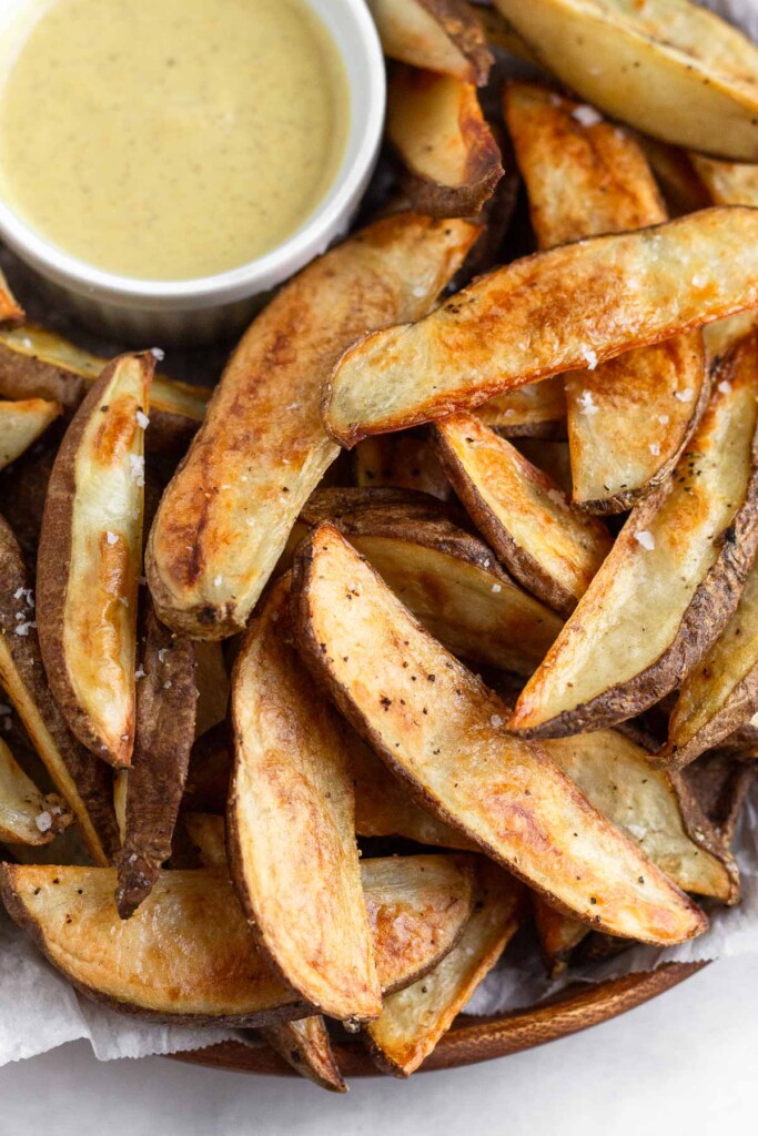 A pile of crispy potato wedges with a ramekin of honey mustard on a plate.