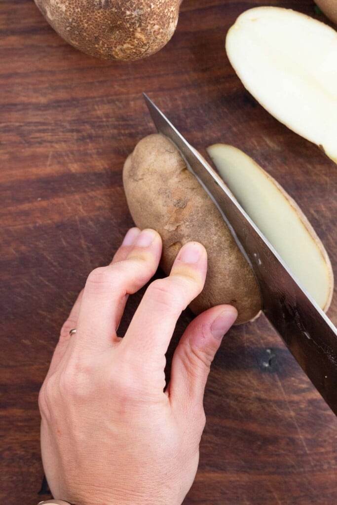 Half of a russet potato on a cutting board being sliced with a knife.