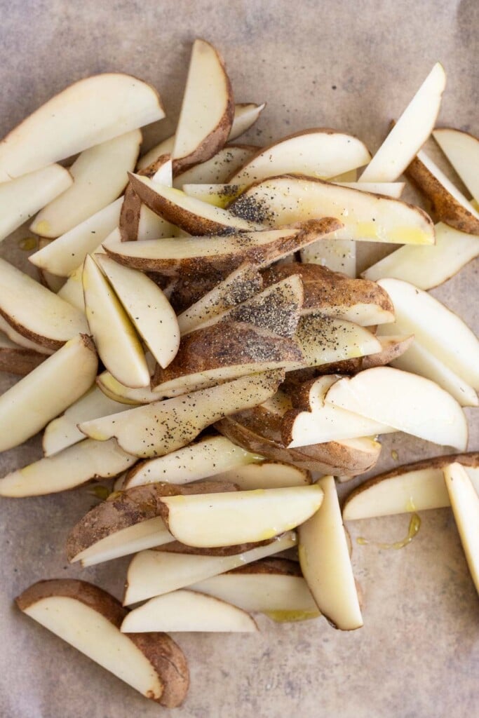 Overhead shot of a pile of raw potatoes wedges with oil, salt, and pepper on them.