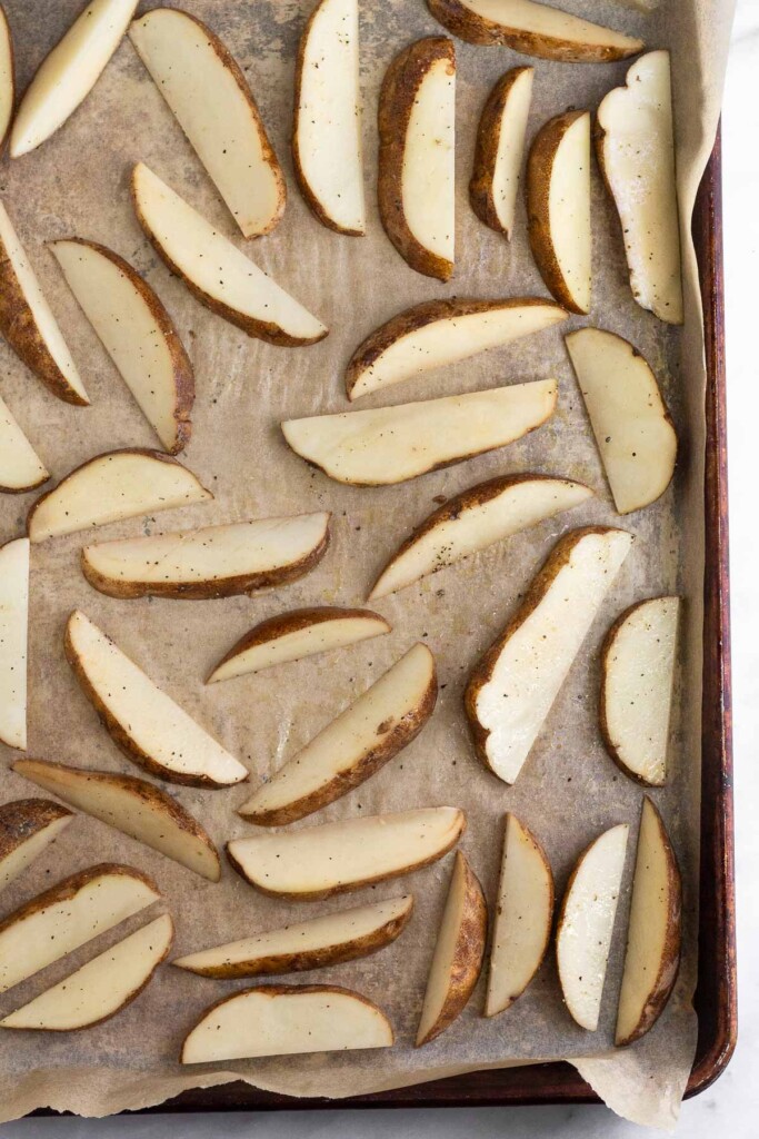 Uncooked potato wedges spread out on a baking sheet lined with parchment paper.