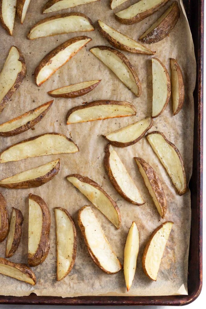 Halfway cooked potato wedges spread out on a baking sheet lined with parchment paper.