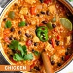 Large pot of chicken fajita soup topped with cilantro, sliced jalapeños, and a lime wedge with a spoon coming out of the pot. The pot is sitting on a striped kitchen towel and around it is a bunch of cilantro and some tortilla chips.