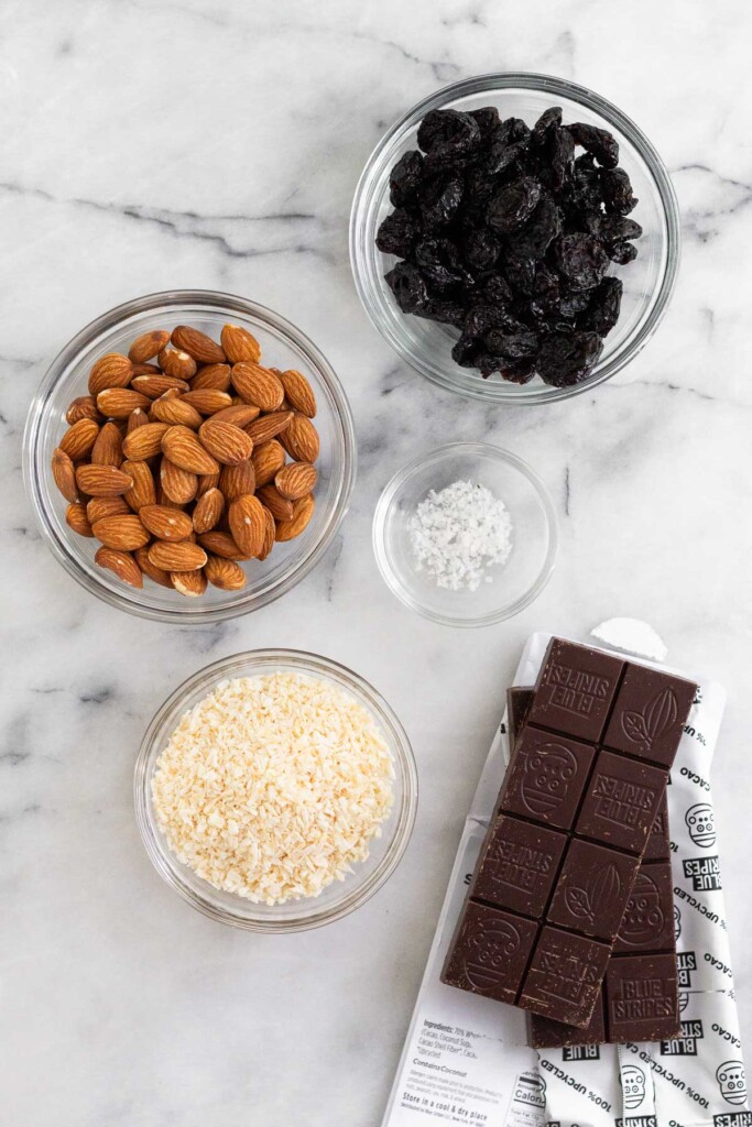 White marble counter with a bowl of dried cherries, two bars of dark chocolate that have been unwrapped, a bowl of shredded coconut, a bowl of sea salt, and a bowl of almonds.