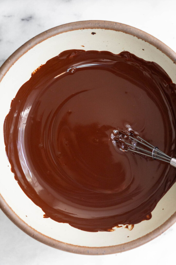 White bowl with melted dark chocolate and a small whisk in it.