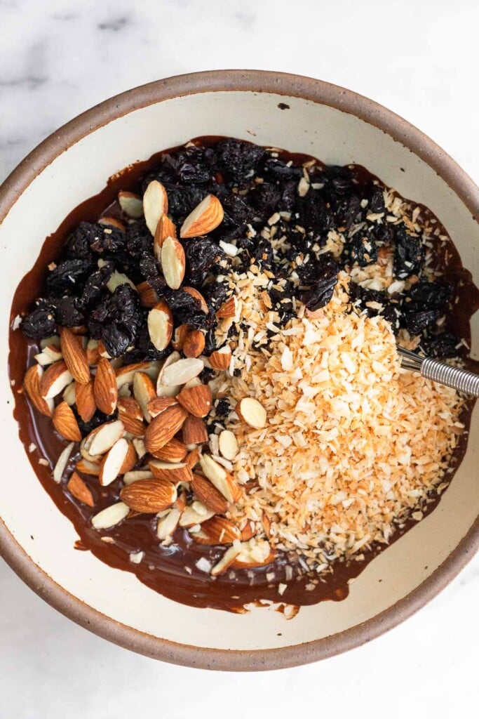 White bowl with chopped dried cherries, chopped almonds, and toasted coconut shreds sitting on top of melted dark chocolate.