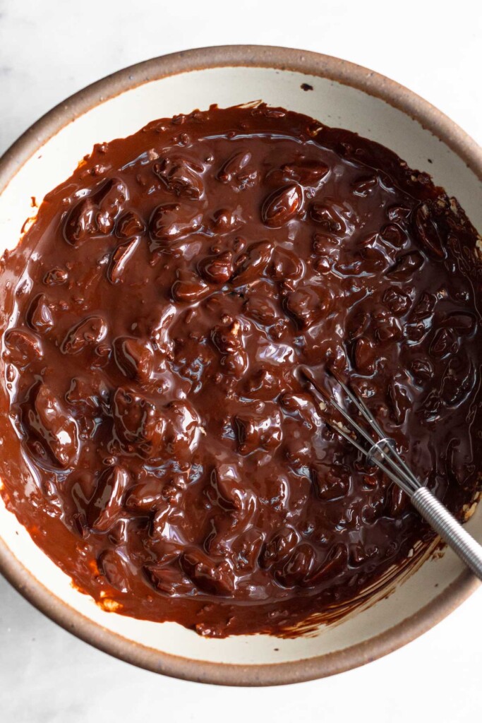 Bowl of melted dark chocolate mixed with nuts and dried fruit. A small whisk is in the bowl.