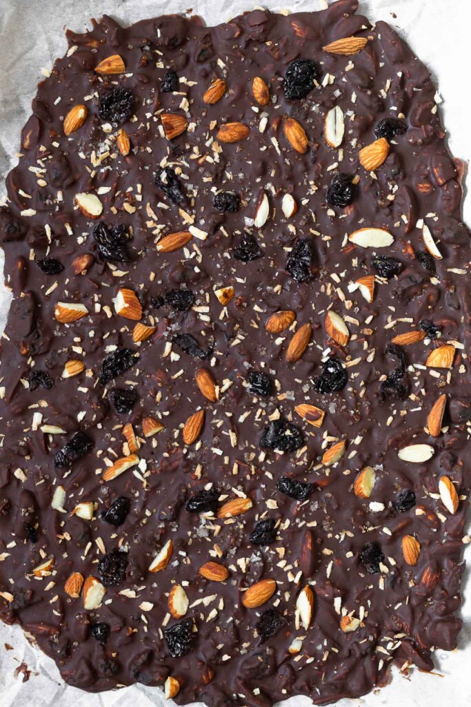 Dark chocolate bark on a baking sheet lined with parchment paper before it is broken into pieces. It has almonds, dried cherries, and coconut in it.
