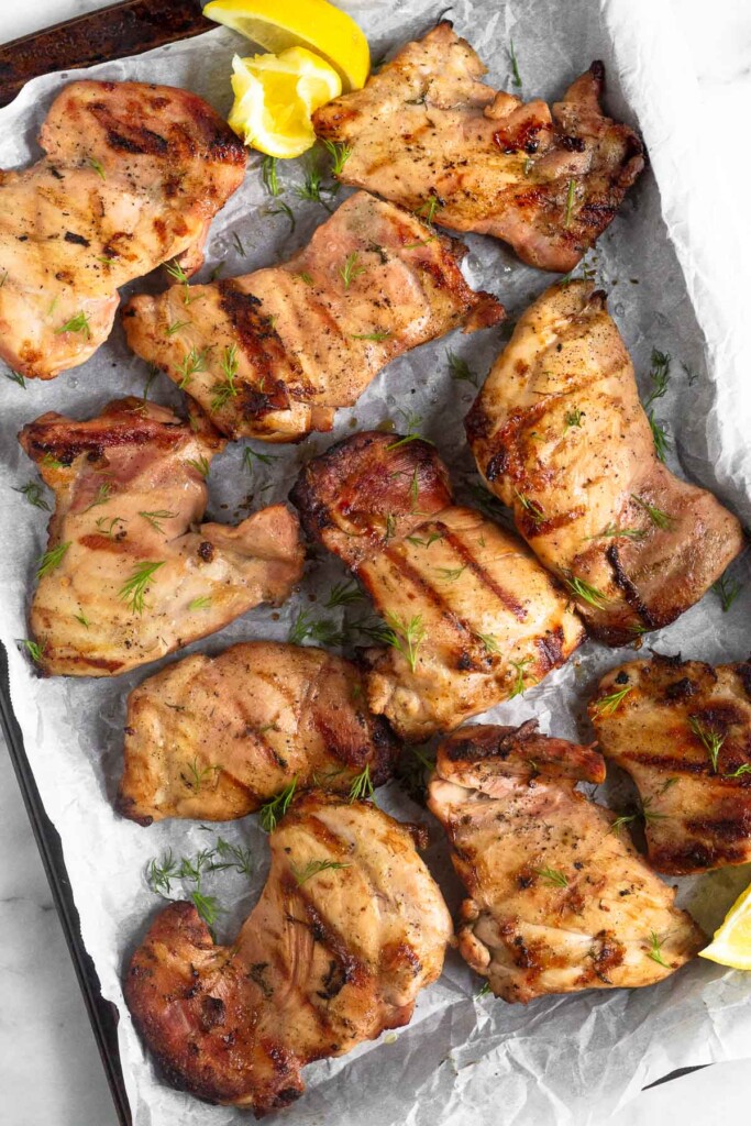 A bunch of pieces of grilled pickle brine chicken on a baking sheet garnished with lemon wedges and fresh dill.