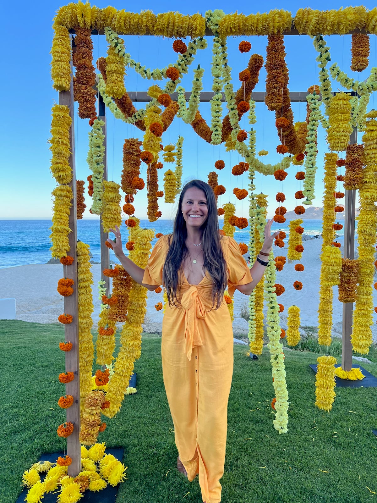 A woman in a yellow dress walking through an arbor decorated with hanging flowers. Behind her is the beach.