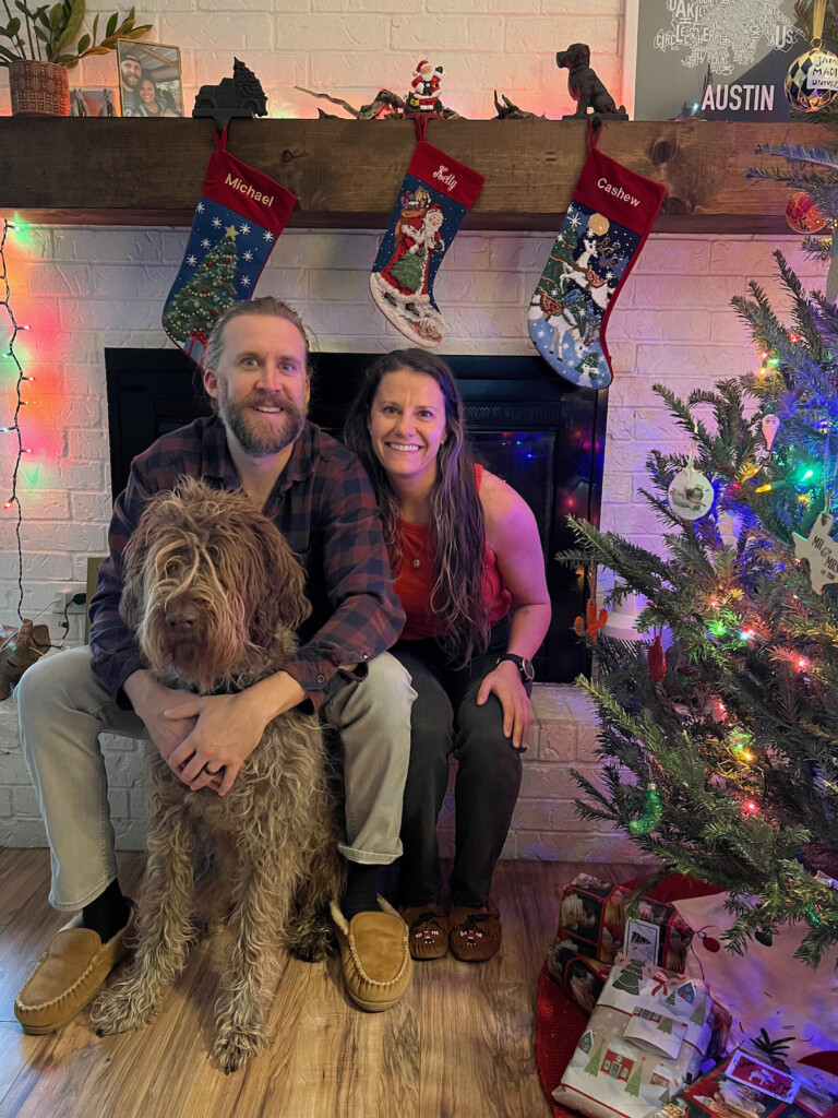 Husband, wife, and their dog sitting in front of a fire place with stockings hanging behind them. A decorated Christmas tree is next to them with the lights on.