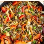 Healthy one pan meal with ground beef, shredded cabbage, bell peppers, and carrots in a large cast iron skillet. It is garnished with green onions. Next to the pan is a bowl of green onions and a bowl of lime wedges.
