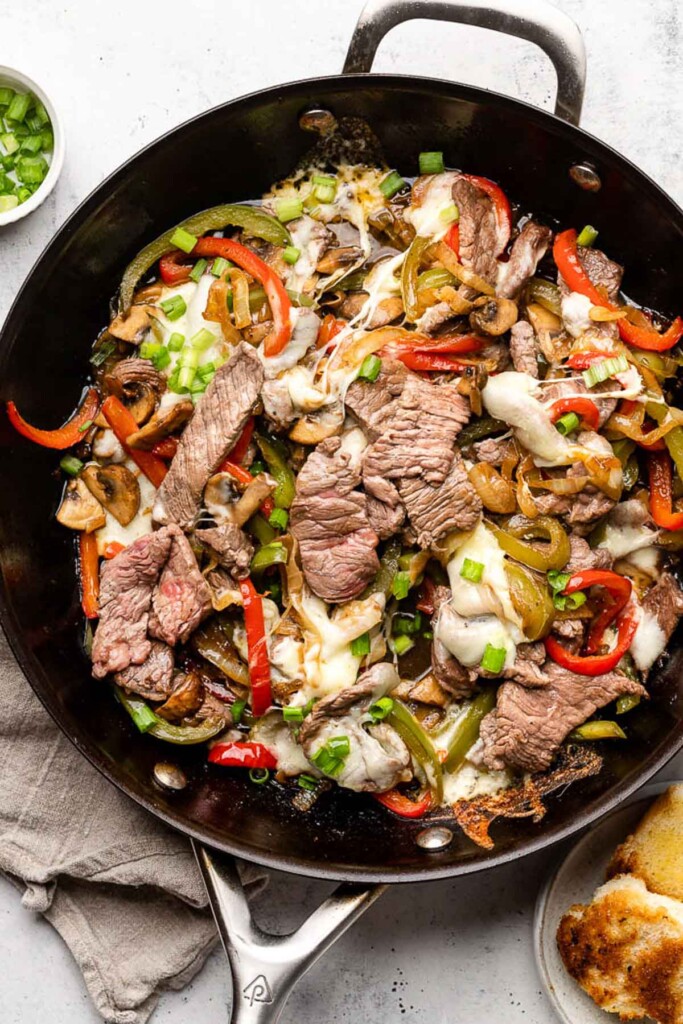 Overhead shot of a skillet with sliced steak, peppers, and onions covered in melted cheese and topped with green onions. Next to the pan is a plate of toasted bread and a bowl of green onions.