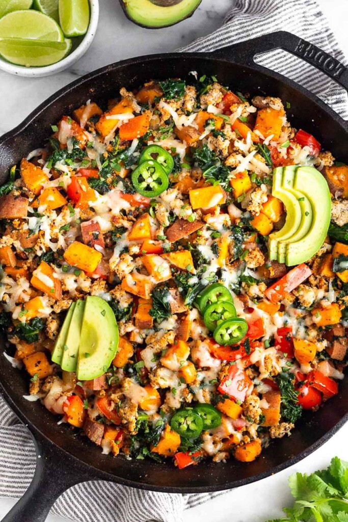 Healthy one pan meal with ground turkey, sweet potatoes, bell peppers, and kale topped with a little bit of melted cheese. It is garnished with avocado and jalapeños. Around the pan is a bowl of lime wedges, half an avocado, and a bunch of cilantro.