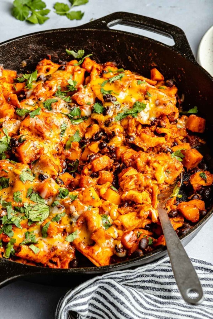 Vegetarian enchilada skillet with black beans and butternut squash topped with melted cheese and garnished with cilantro. A spoon is in the skillet.