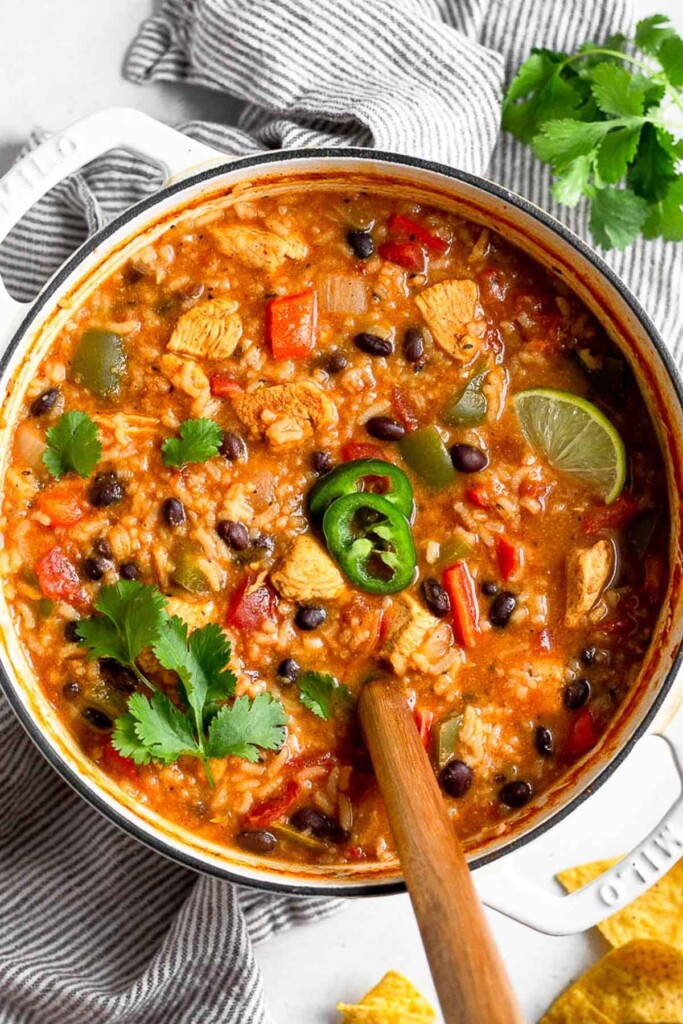 Large pot of soup with chicken, rice, black beans, and peppers. It is garnished with cilantro, sliced jalapeños, and a lime wedge. Next to the pot is a bunch of cilantro and some tortilla chips. A spoon is in the pot.