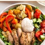 High protein lunch recipe in a bowl with greens, roasted veggies, sliced chicken thighs, a tomato and cucumber salad, and a yogurt sauce on top. Around the bowl is a sheet pan of more roasted veggies and a bowl of yogurt sauce.
