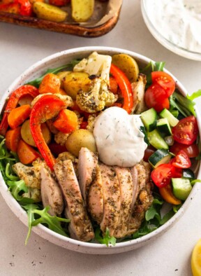 High protein lunch recipe in a bowl with greens, roasted veggies, sliced chicken thighs, a tomato and cucumber salad, and a yogurt sauce on top. Around the bowl is a sheet pan of more roasted veggies and a bowl of yogurt sauce.