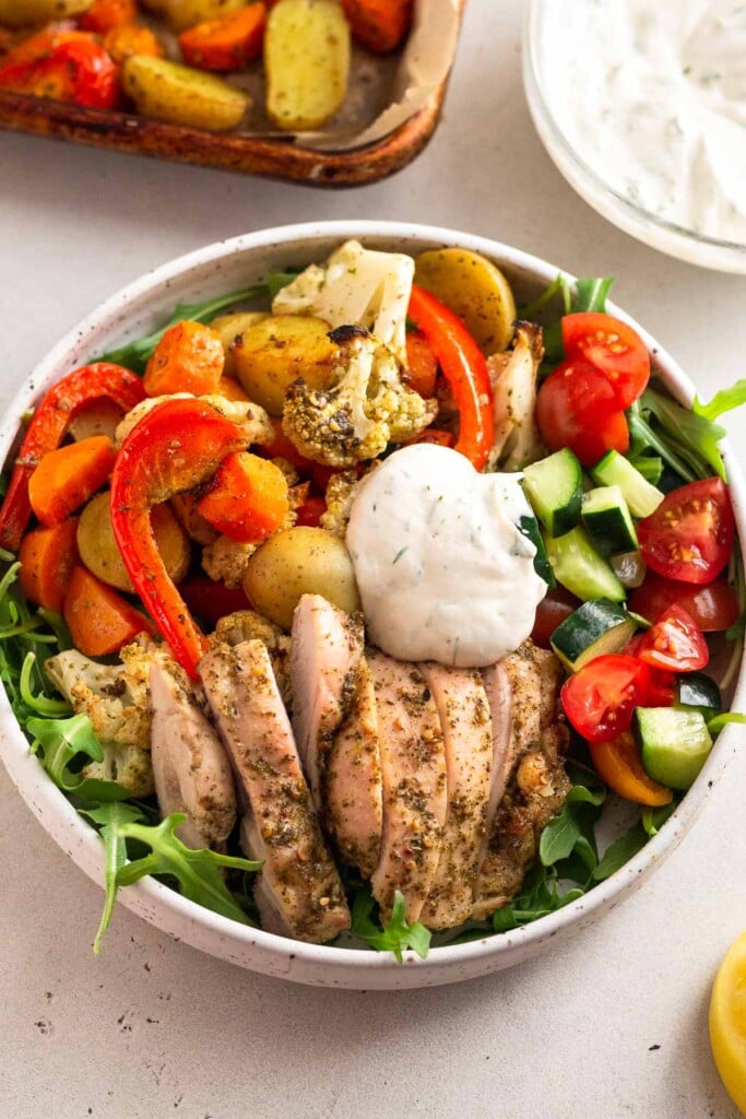 High protein lunch recipe in a bowl with greens, roasted veggies, sliced chicken thighs, a tomato and cucumber salad, and a yogurt sauce on top. Around the bowl is a sheet pan of more roasted veggies and a bowl of yogurt sauce.