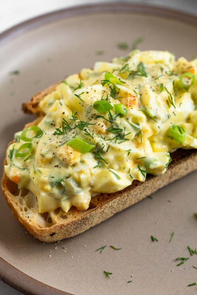 A close up of healthy egg salad on a piece of toast on a brown plate. It is topped with sliced green onions, dill, and black pepper.