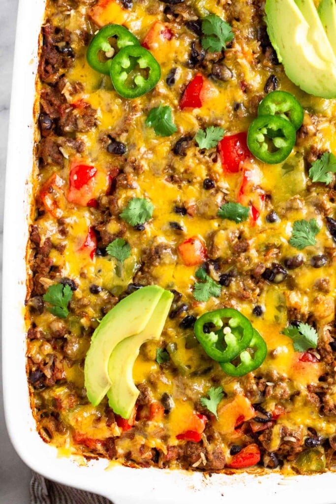 Beef burrito casserole topped with melted cheese in a large casserole dish. It is topped with sliced avocado, sliced jalapeño, and cilantro.
