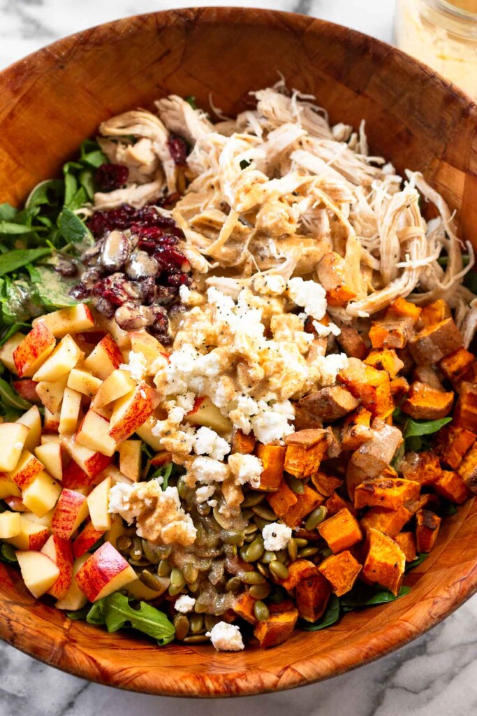 Large wooden salad bowl with shredded chicken, roasted sweet potatoes, pepitas, diced apple, dried cranberries, and goat cheese over mixed greens. A creamy dressing is poured overtop.