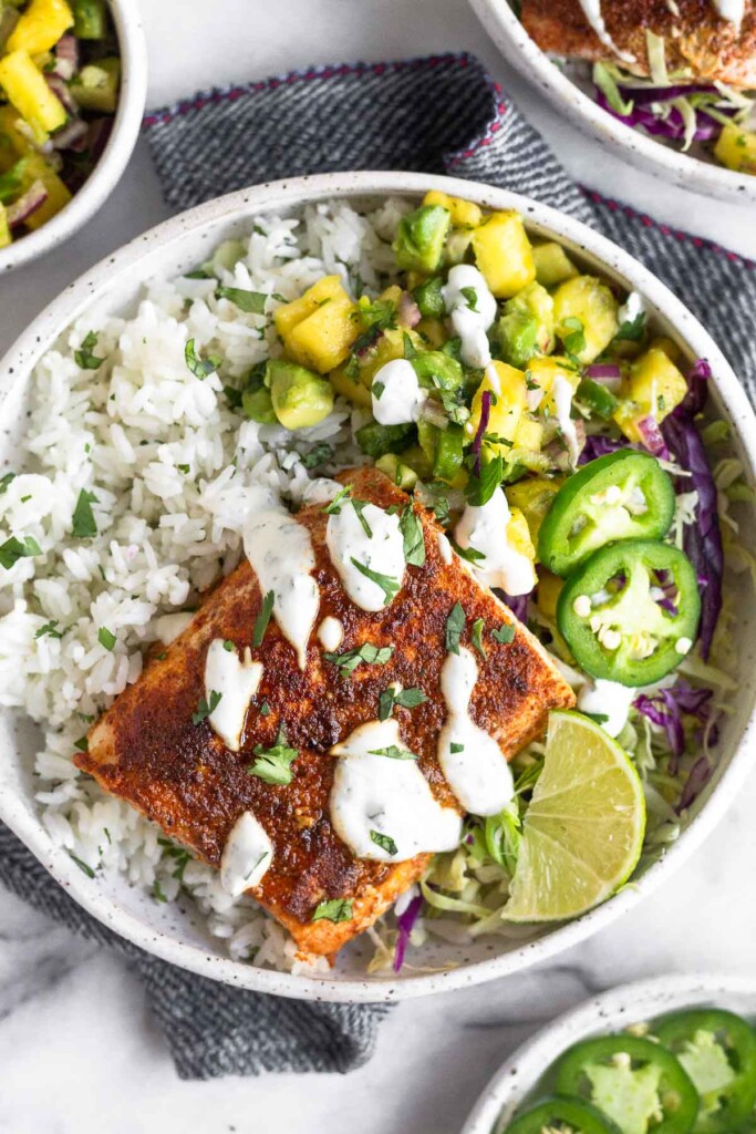 Fish taco bowl with cilantro white rice, pineapple salsa, cabbage, seasoned fish, and white sauce in a bowl. Around it is a plate of jalapeños, a bowl of salsa, and another fish taco bowl.
