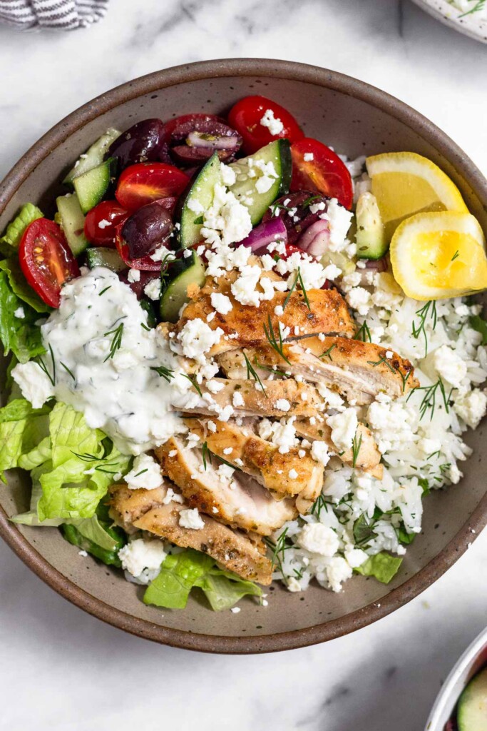 High protein lunch recipe in a bowl with chopped romaine, tomato cucumber salad, herby rice, sliced chicken thighs, tzatziki, and feta and garnished with dill and 2 lemon wedges.