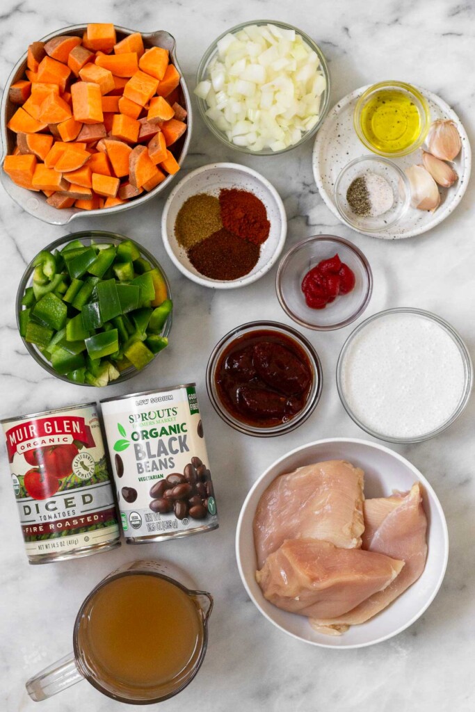 White marble counter with a bowl of diced onions, a small plate with 3 cloves of garlic, a bowl of olive oil, and a bowl of salt and pepper, a bowl of tomato paste, a bowl of coconut milk, a bowl of raw chicken breasts, a bowl of chipotle peppers in adobo sauce, a cup of chicken broth, a can of black beans, a can of fire roasted diced tomatoes, a bowl of diced green bell peppers, a bowl of spices, and a bowl of diced sweet potatoes.