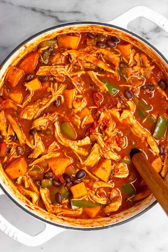 Overhead shot of chicken and sweet potato soup in a large white dutch oven with a spoon in it on a marble countertop.