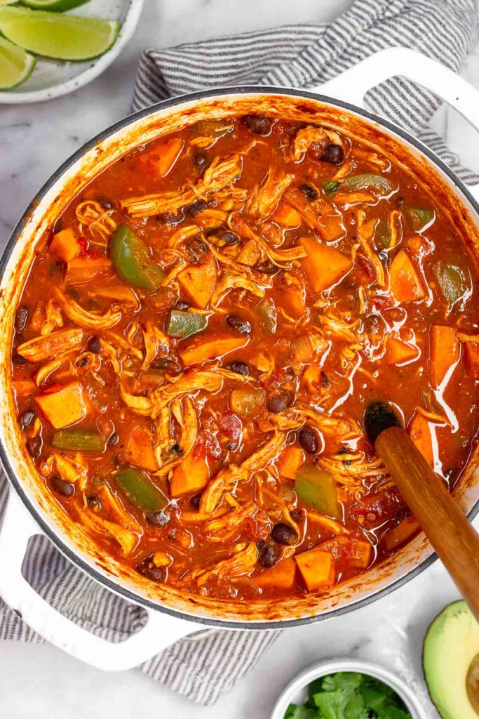 Chicken sweet potato soup in a large white dutch oven with a spoon in it. The pot is sitting on a striped towel with half an avocado, a bowl of cilantro, and a plate of lime wedges around it.